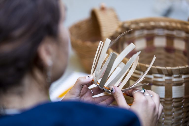 Over the shoulder photo of a woman weaving a basket from strips Black Ash. 