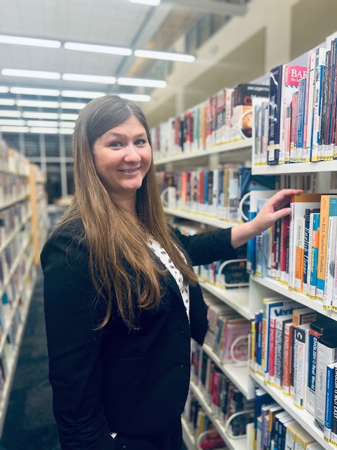 Michelle Gibbons, a white woman with long light brown hair, stands smiling in the aisle of a library and reaches for a book on a shelf.