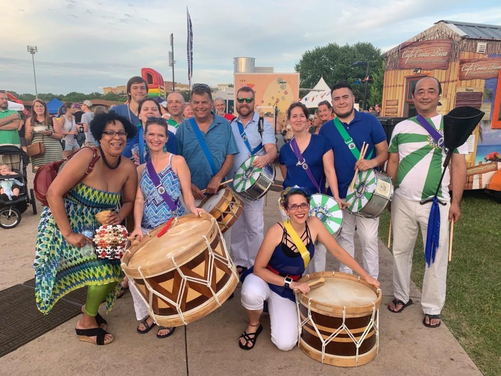 Diverse group of 11 people smiling outdoors holding various drums with green and white rays on them, some with matching shirts.