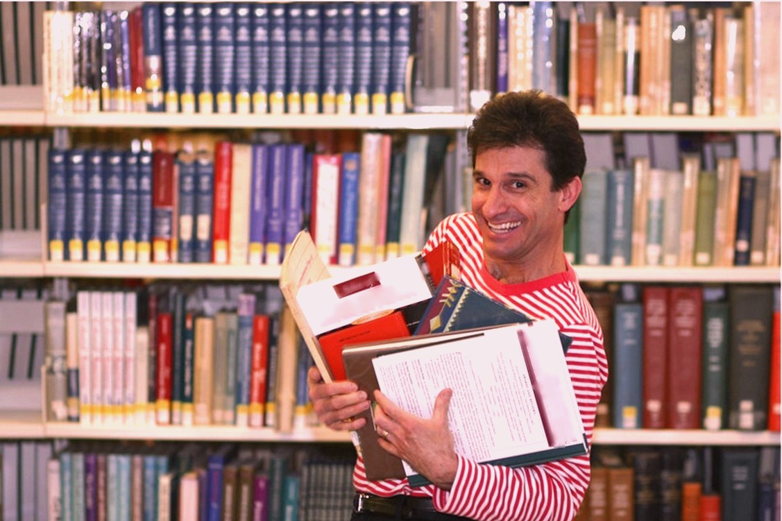 Chris Fascione, a white man with short dark hair wearing a shirt with thin horizontal red and white stripes, stands smiling and holding heaping armfuls of books with library bookshelves in the background.