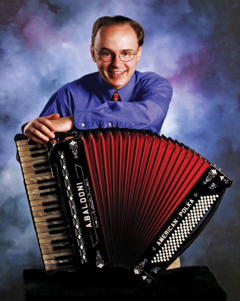 Mike Schneider, a smiling white man wearing glasses and a blue button down shirt, poses with his accordion against a blue and purple background.