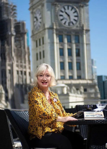 Veronica Hinke, a white woman with pale blond medium length hair wearing a vibrant yellow blouse with an elegant black floral pattern, sits smiling at an outdoor cafe table. The table holds a typewriter, a copy of her book about dining on the Titanic, and a crystal cocktail glass. A clock tower stands in the background. 