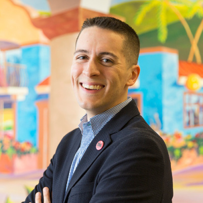 Sergio González, a light-skinned Mexican man with short dark hair smiles charismatically. He is wearing a dark gray suit jacket and blue checkered shirt and stands against a brightly colored mural in the background.